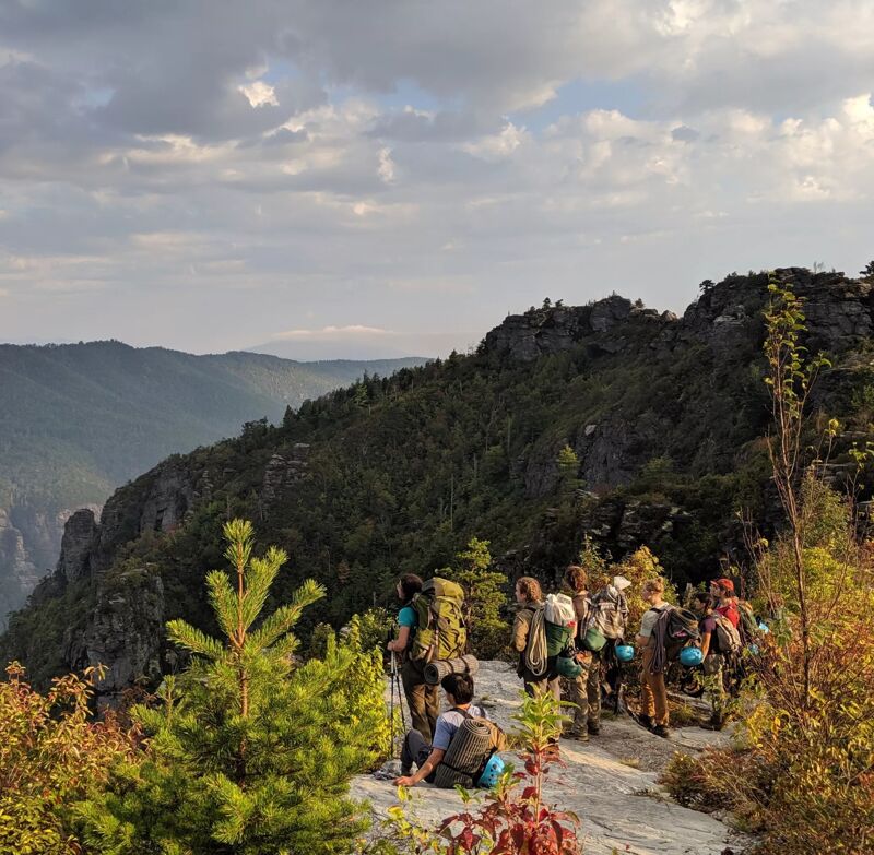 A group of hikers is standing on a rocky outcrop, taking in the view of a mountainous landscape. The sky is filled with clouds, and the sun is shining through, creating a dramatic effect. The hikers are wearing backpacks and hiking gear, and they appear to be enjoying their time in nature. The vegetation is lush and green, and the overall scene is one of peace and tranquility.